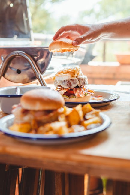 A close-up view of a person's hand assembling a burger, placing the top bun onto a layered filling that includes lettuce, tomato, and a beef patty. In the foreground, a plate of crispy, golden-brown f