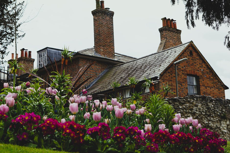 A peaceful garden scene featuring a well-maintained grassy lawn bordered by lush green shrubs and flowering plants with pink blossoms. On the left side, a wooden garden bench is situated among dense f